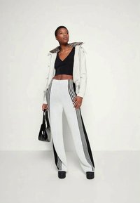 Jovem mulher com um casaco branco com colarinho de pelo, top preto cropped, calças brancas com riscas pretas, sapatos de plataforma pretos, segurando uma bolsa preta.