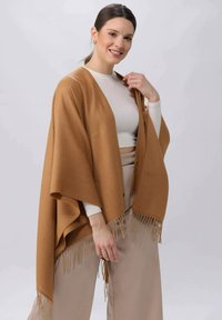 Brauner Poncho mit Fransenbesatz, aus einem weichen, strukturierten Stoff gefertigt. Verfügt über ein lockeres, offenes Design mit weit geschnittenen Ärmeln. Leicht und fällt elegant.