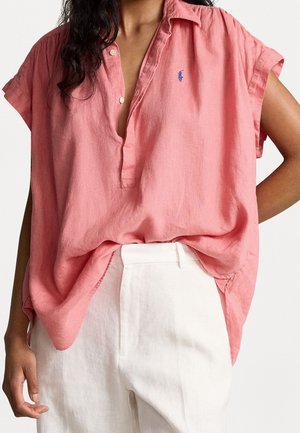 Mujer con una camisa rosa de cuello, suelta y de manga corta, parcialmente metida en pantalones blancos de talle alto, de pie con un brazo doblado.