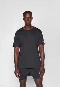 Schwarzes Baumwoll-T-Shirt mit kurzen Ärmeln und rundem Ausschnitt. Mit einem kleinen Logoakzent auf dem linken Ärmel. Glatte Textur.