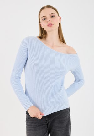 Junge Frau in einem hellblauen, schulterfreien Strickpullover und schwarzer Jeans, die mit einer Hand in der Tasche vor einem schlichten weißen Hintergrund posiert.