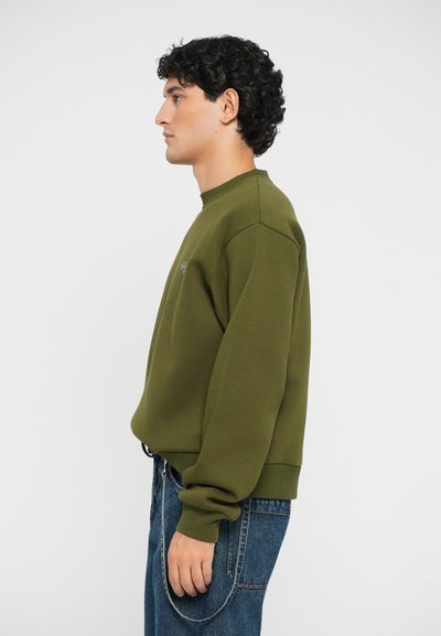 Felpa verde oliva con collo a giro, polsini a coste e vestibilità rilassata, abbinata a jeans blu. Design minimalista senza decorazioni.