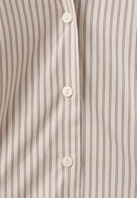 Gros plan sur un tissu beige à fines rayures verticales marron et trois boutons blancs le long d'une patte de boutonnage de chemise.