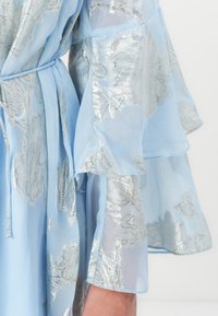 Robe transparente azul claro com padrões florais metálicos, mangas onduladas e cintura atada. Textura suave com um design elegante e fluido.