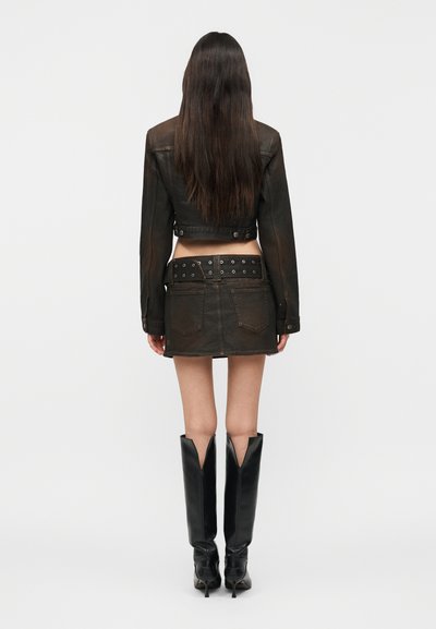 Femme de dos portant une veste en cuir noire courte, une mini-jupe noire avec ceinture et des bottes noires à talons montant jusqu'aux genoux sur un fond uni.