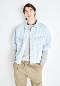 Chaqueta de denim azul claro con dos bolsillos en el pecho, botones plateados y un corte relajado, llevada sobre un suéter gris de canalé y pantalones caqui.