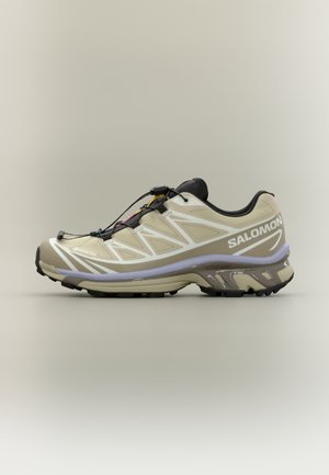 Hellbeiger Trailrunningschuh mit schwarzen Akzenten, Mesh-Obermaterial, gepolsterter Sohle, elastischem Schnürsystem und strukturiertem Gummiprofil.