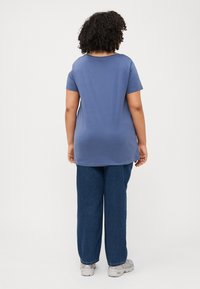 Una persona con cabello rizado está de espaldas, vistiendo una camiseta azul y unos jeans azules holgados, frente a un fondo blanco liso.