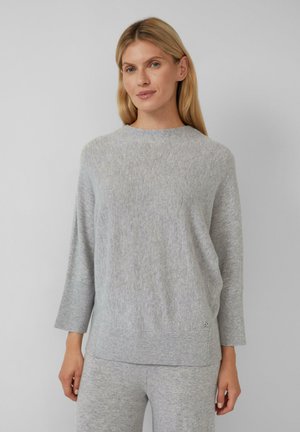 Blonde Frau trägt einen hellgrauen Strickpullover mit 3/4-Ärmeln und passende graue Strickhosen, steht vor einem schlichten Hintergrund.