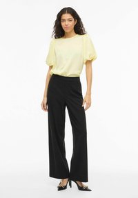 Blouse jaune transparente avec manches bouffantes, associée à un pantalon noir à jambes larges. Le mannequin porte des talons noirs avec un accent doré. Fond simple.