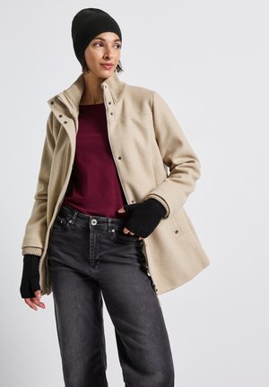 Giacca in pile beige con colletto alto e bottoni a pressione, abbinata a una camicia bordeaux, jeans in denim neri e guanti senza dita neri.