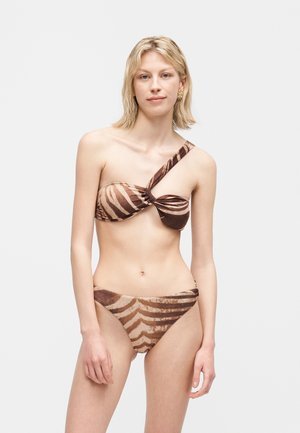 Νέα γυναίκα που φοράει καφέ μπικίνι με ένα ώμο και μοτίβο ζέβρας, με σκουλαρίκια, στέκεται μπροστά σε απλό λευκό φόντο.