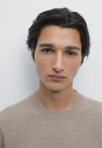 Młody mężczyzna z ciemnymi, falującymi włosami ma na sobie jasnobrązowy, teksturowany sweter. Ma neutralny wyraz twarzy i jednolite tło.