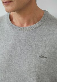 Jasnoszary sweter z okrągłym dekoltem, wykonany z miękkiego materiału. Posiada ribbowany dekolt oraz małe logo w ciemnoszarym kolorze na piersi.
