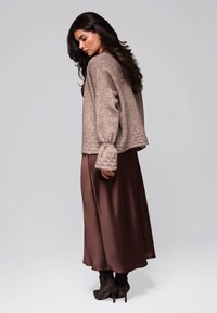 Brauner strukturierter Pullover mit gepufften Ärmeln, kombiniert mit einem braunen Satin-Maxikleid und schwarzen High-Heels, vor einem neutralen Hintergrund.