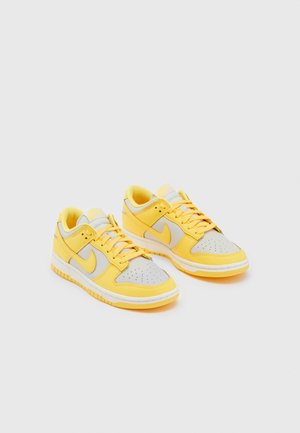 Para niskich butów sportowych w jasnoszarym i jaskrawożółtym kolorze z perforowanym noskiem, sznurowanym fasonem i gumowymi podeszwami na jednolitym tle.