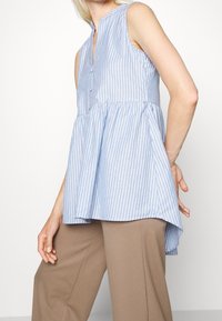 Haut sans manches à rayures bleu clair avec une patte de boutonnage, taille froncée et un ourlet arrière plus long, fabriqué dans un tissu léger.