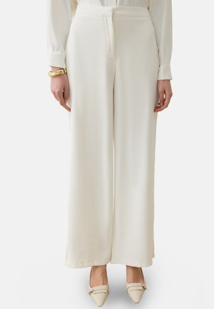 Donna che indossa pantaloni a vita alta e gamba larga color avorio, scarpe crema con punta affilata e un braccialetto d'oro al polso sinistro.