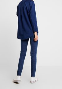 Marineblauw longsleeve shirt met knoopmanchetten, afgeronde zoom, gecombineerd met aansluitende spijkerbroek en witte sneakers. De stof lijkt soepel en rekbaar te zijn.