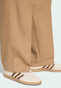 Pantalones de pierna ancha beige con textura acanalada vertical, combinados con zapatillas de rayas crema y marrón con suela de goma.