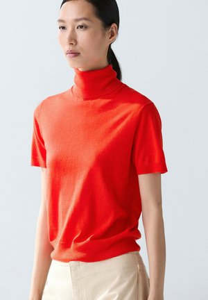 Donna con capelli neri legati indossando un maglione dolcevita rosso brillante a maniche corte e pantaloni beige, in piedi davanti a uno sfondo chiaro uniforme.