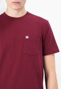 Bordeaux t-shirt lavet af bomuld med korte ærmer, rund halsudskæring og en brystlomme, der har et lille hvidt GAP-etiket.