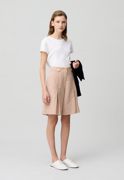 Camiseta blanca de manga corta combinada con pantalones cortos a rayas de longitud hasta la rodilla en beige y blanco. Llevado con zapatos blancos y sosteniendo una chaqueta negra.