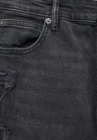 Detailní záběr na černé džíny z denimového materiálu, ukazující přední kapsu, poutko na opasek, kovové nýty a roztržený detail na látce.