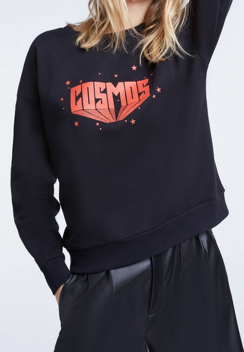 Femme portant un sweat-shirt noir avec le texte rouge "COSMOS" et des motifs d'étoiles, associé à un pantalon en cuir noir, fond clair.