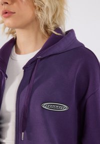 Ljubičasta dukserica na zip s prednjim džepom, podesivim kapuljačom i crnom logotipom na lijevoj strani prsnog predjela. Mekana pamučna tkanina.