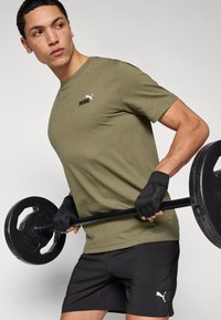 Λαδί πράσινο t-shirt PUMA με λευκό λογότυπο, σε συνδυασμό με μαύρο σορτς. Το άτομο κρατά μια μαύρη μπάρα με γάντια, δείχνοντας ένα περιβάλλον γυμναστικής.