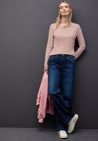 Jonge vrouw staat tegen een grijze muur, gekleed in een lichtroze lange mouw top, donkerblauwe jeans, witte sneakers en houdt een roze jas in haar hand.