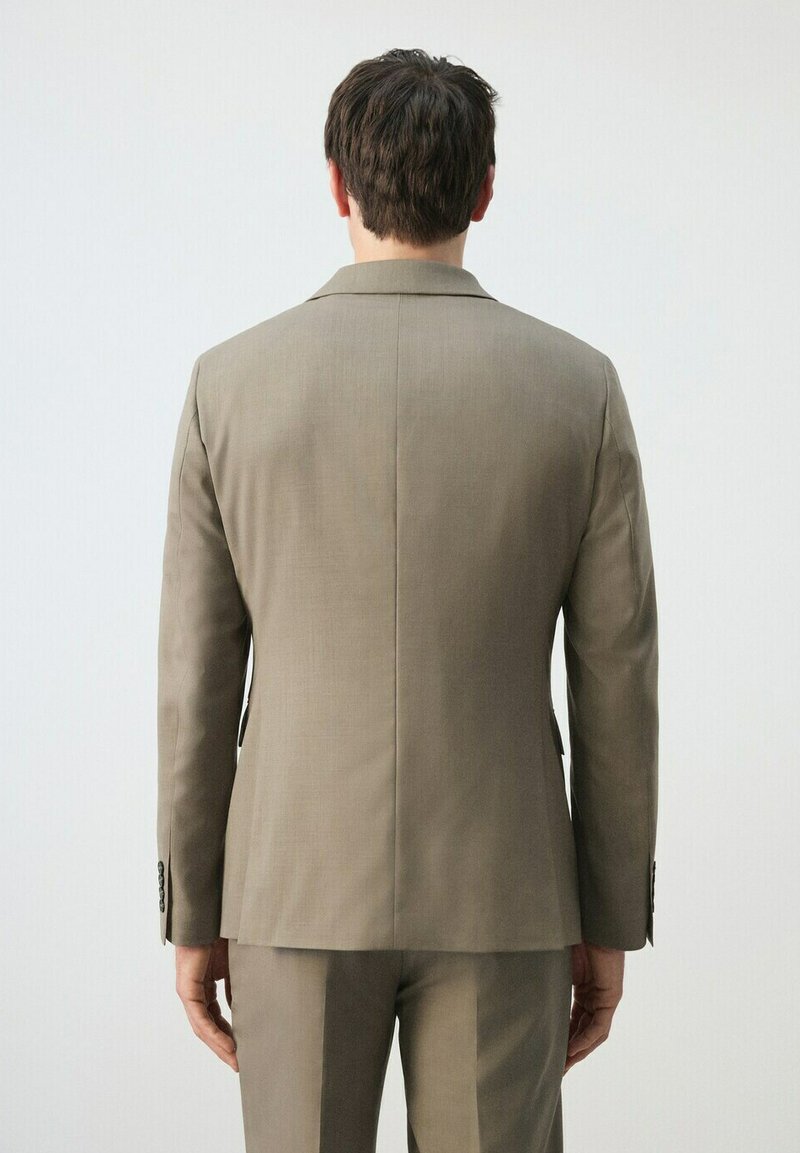 Man die met de rug naar de kijker staat, gekleed in een lichtbruin colbert en bijpassende broek tegen een effen witte achtergrond.