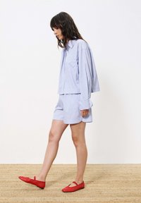 Blauw en wit gestreept shirt met lange mouwen, gecombineerd met bijpassende shorts. De outfit wordt aangevuld met felrode ballerina's en een vloerkleed van natuurlijke vezels.