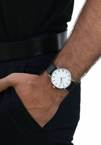 Eine Nahaufnahme des Handgelenks eines Mannes, der eine schwarze Armbanduhr mit weißem Ziffernblatt trägt, während seine Hand in einer Tasche ruht, betont durch einen schwarzen Gürtel.