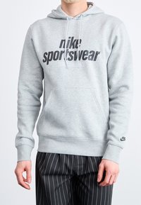 Gray Nike sportswear hoodie with kangaroo pocket, drawstring hood, and large printed logo. Ribbed cuffs and hem. Worn with pinstriped pants.