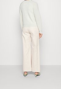 Kvinna som bär en krämfärgad stickad tröja, vid-beinade beige byxor och ljusgröna kitten heels som står mot en enkel bakgrund.