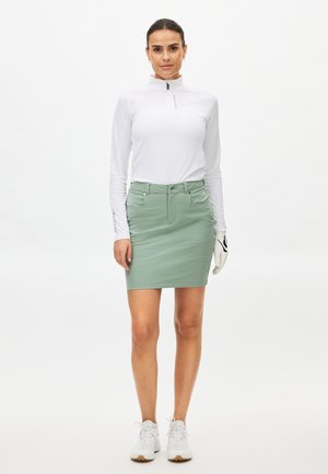 Weißes Langarmshirt mit Reißverschluss-Kragen, kombiniert mit einem hellgrünen Rock und weißen Sportschuhen. Handschuh in der rechten Hand gehalten. Schlichtes Design.