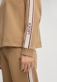 Felpa color sabbia con maniche lunghe, caratterizzata da un design a righe bordeaux e crema e la scritta "JOOP!" lungo la manica. Tessuto morbido.