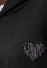 Hoodie nera con un ricamo a forma di cuore di colore viola scuro e texture, accompagnato da un testo stilizzato, posizionato sulla parte sinistra del petto.