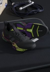 Paire de chaussures de sport noires avec des accents verts néon et violets placées sur une surface sombre, avec une corde à sauter en arrière-plan.