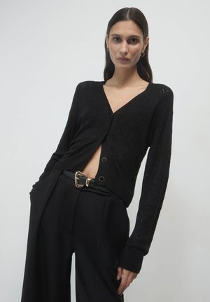 Schwarzer Strickcardigan mit V-Ausschnitt, ausgestattet mit einem strukturierten Muster und zwei vorderen Knöpfen. Kombiniert mit hochgeschnittenen schwarzen Hosen und einem Gürtel.