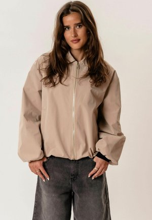 Jauna moteris su ilgais rudais plaukais, vilkinti smėlio spalvos striukę su užtrauktuku ir tamsiai pilkas kelnes, rankos kišenėse, priešais paprastą šviesią foną.