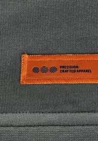 Grijze stoffen label met oranje rand, voorzien van de tekst "Precisie-Vakmanschap Kleding" en drie verschillende patronen in donkerblauw.