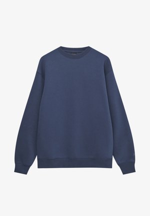 Person trägt einen weiten navyblauen Pullover, weite cremefarbene Hosen mit Gesäßtaschen und weiße Sneaker, steht mit dem Rücken zur Kamera vor einem schlichten weißen Hintergrund.