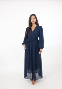 Vrouw staand in een marineblauwe maxi-jurk met lange mouwen, een V-hals en een geplooide taille, met doorzichtige sandaalbandjes op een witte achtergrond.