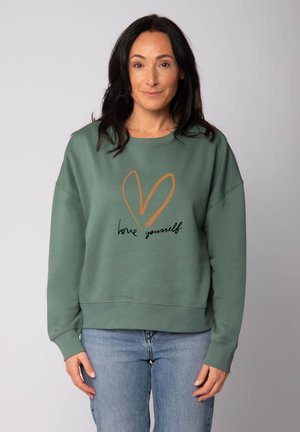 Sweatshirt vert à manches longues, coupe décontractée, avec un motif en forme de cœur en brun et un texte noir disant "aime-toi" à l'avant.