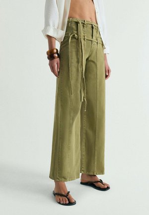 Persona que lleva pantalones verde oliva de talle alto y pierna ancha con largas cintas de tela para el cinturón, combinados con chanclas negras con cuentas y pulseras de madera.