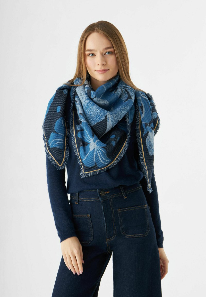 Écharpe bleue à motifs, fabriquée en tissu doux, avec des motifs floraux et des bords effilochés, drapée élégamment autour des épaules du modèle.