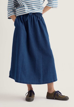 Person mit blau-weiß gestreiftem Oberteil, langem blauen Jeansrock mit Kordelzug in der Taille und olivgrünen Sneakers steht mit den Händen in den Rocktaschen.
